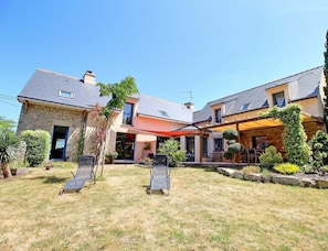 Een tweelaags huis met een overdekt terras, een gazon met twee loungestoelen en diverse planten en bomen.