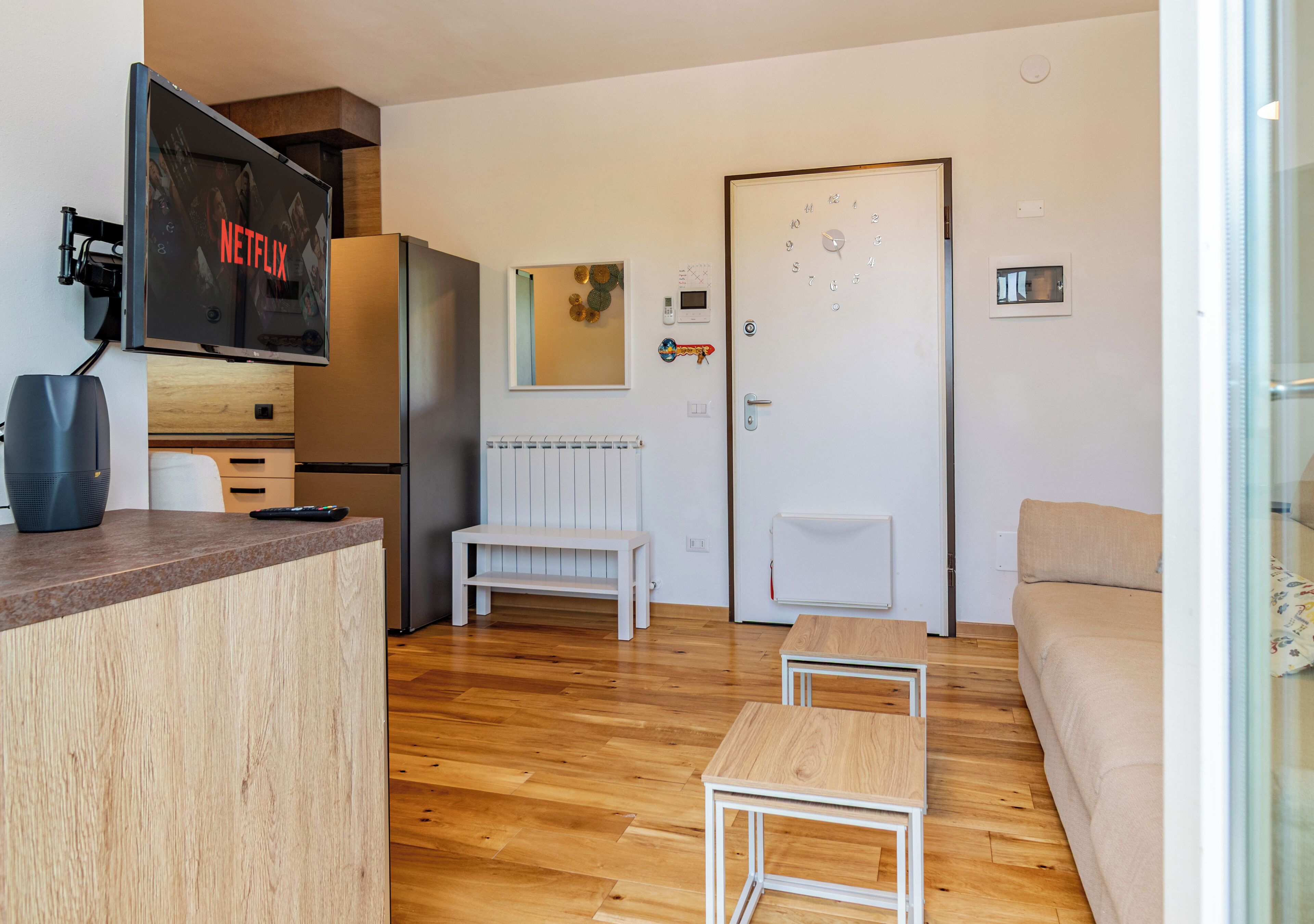 A modern living room with a wooden floor, a white sofa, a small white table, and a TV mounted on the wall displaying 'NETFLIX'.