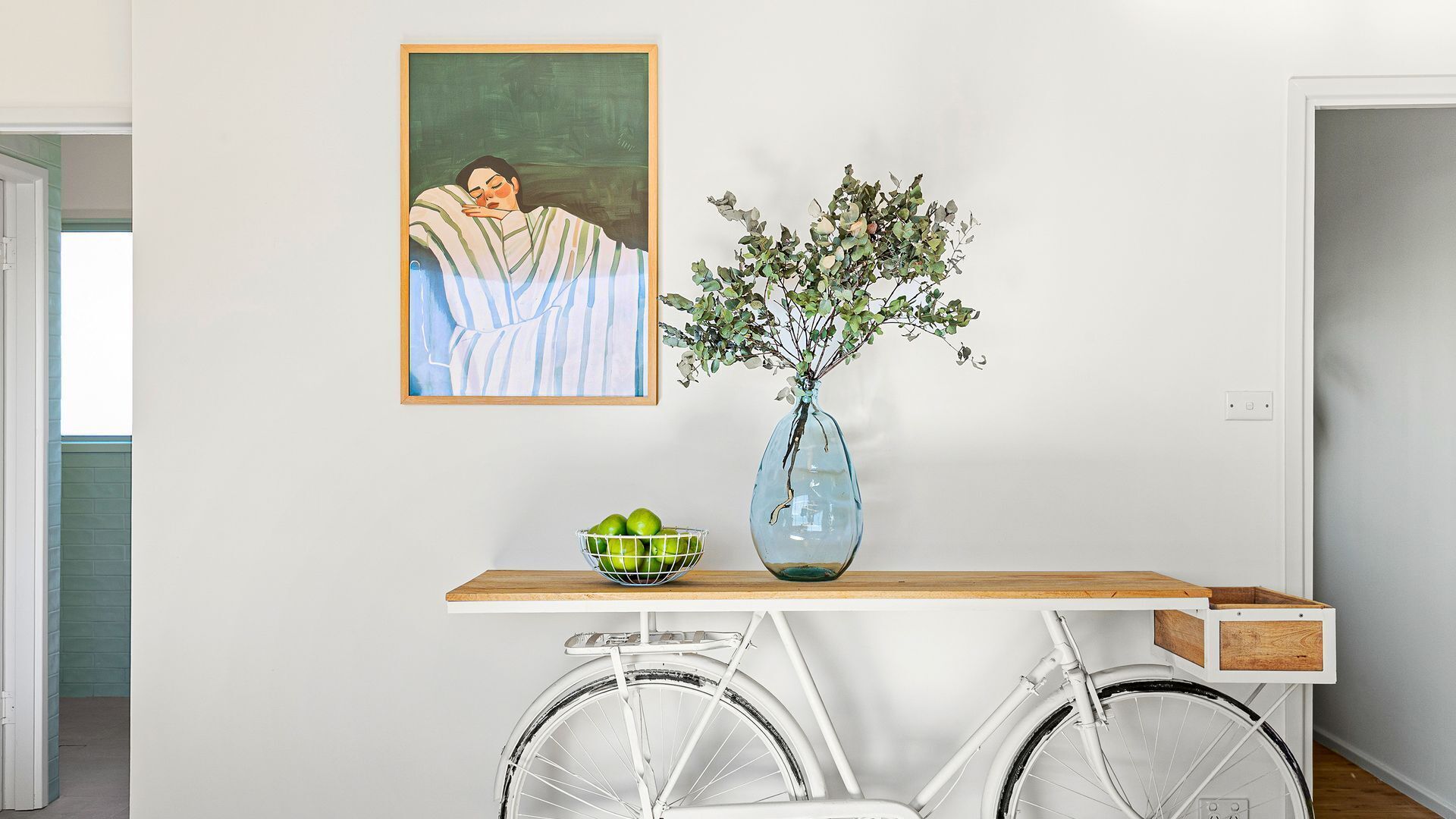Une pièce avec un mur blanc, un tableau encadré, un vase avec des feuilles vertes, un bol de pommes vertes et un vélo blanc.