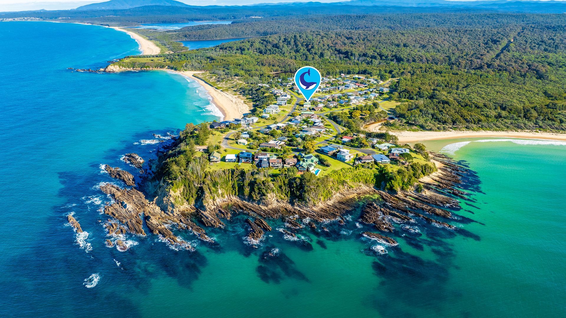 Vue aérienne d’une zone résidentielle côtière, avec des maisons, une plage et un paysage boisé.