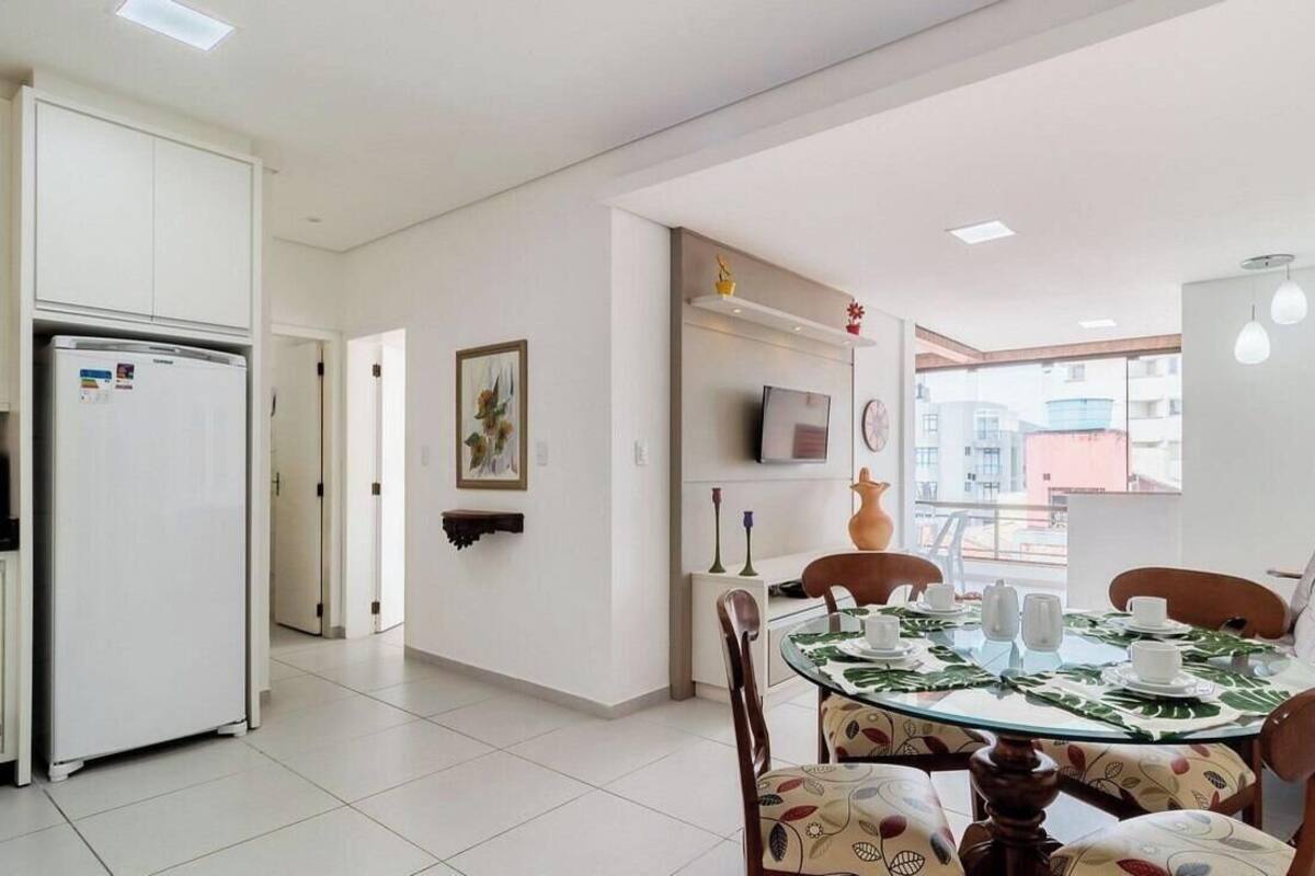 A modern dining area with a glass table, chairs with patterned cushions, a refrigerator, a TV on a shelf, and a view of buildings outside.