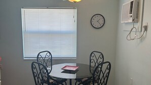 Un coin repas comprenant une table et des chaises, un ventilateur de plafond, une horloge et une fenêtre avec des stores.