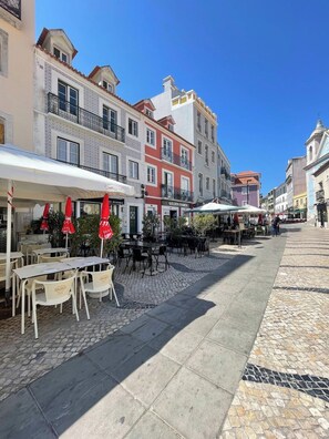 A street with outdoor seating, buildings with balconies, and a clear sky. - Tagus River Apartment *Cacilhas, 10min from Lisbon (Almada)
