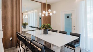 A modern dining area with a large white table, black upholstered chairs, a potted plant, and a chandelier. - Family 5BR Windsor Island Princess Toy Story Rooms (Davenport)