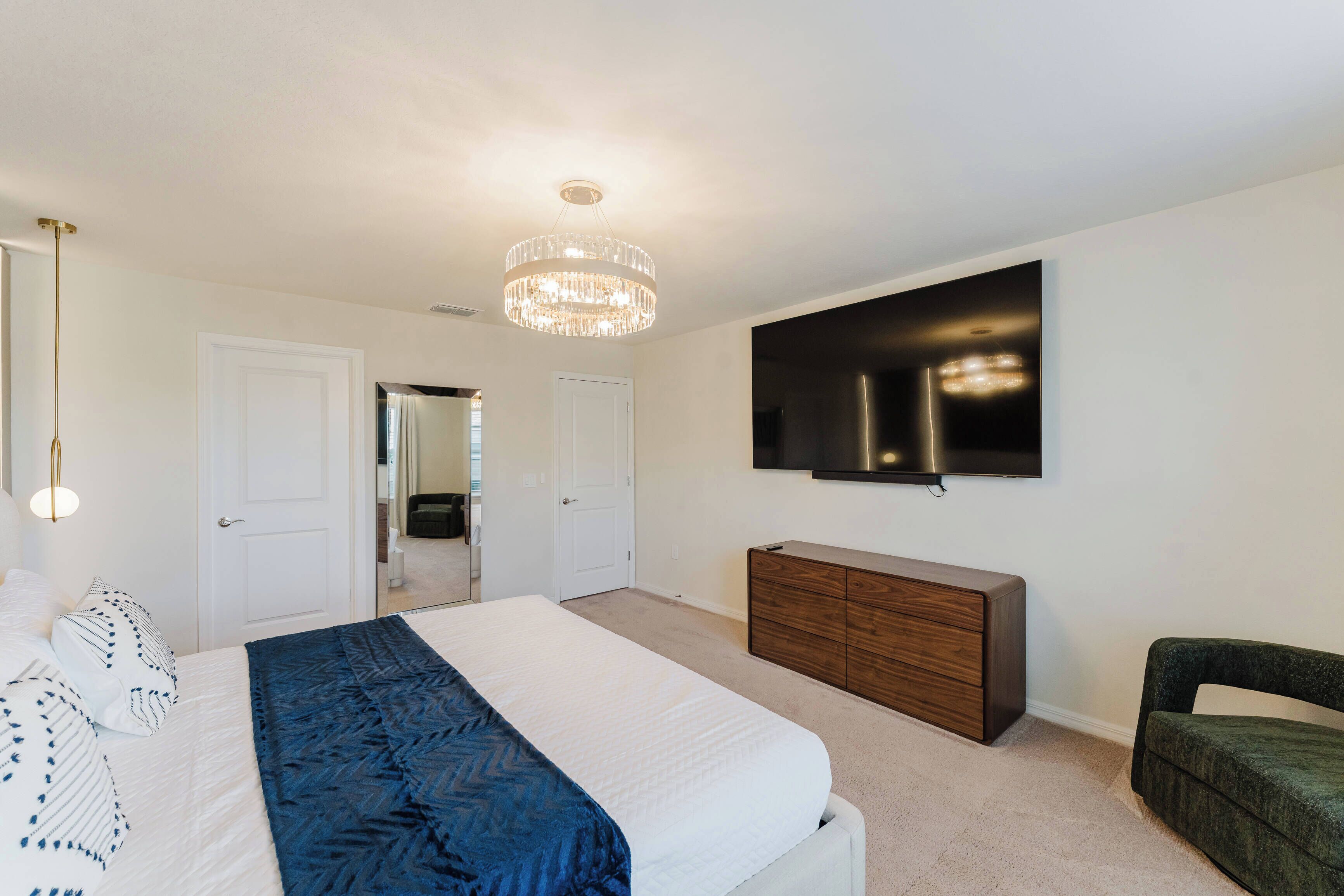 Een slaapkamer met een groot bed, een houten dressoir, een aan de muur bevestigde televisie en een kroonluchter.