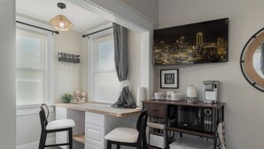 A compact kitchen with a built-in microwave, coffee maker, and a small refrigerator. There are two chairs and a window with blinds. - "KOZY KOTTAGE" Near Downtown! 2025 Remodel, Sleeps 8 Comfortably, Games & More! (Denison)
