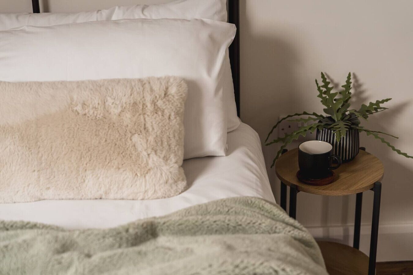 A bed with white bedding and a green throw blanket, a small round wooden side table with a black mug and a potted plant.