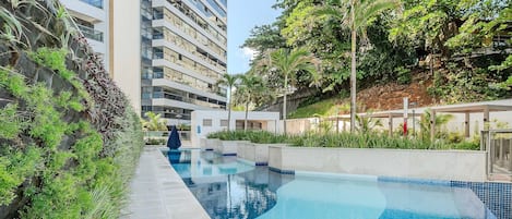 A modern hotel with a swimming pool, surrounded by lush greenery and a multi-story building.