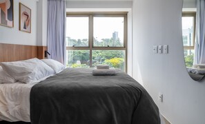 A neatly made bed with white and gray linens, a wooden headboard, and a window with a view of greenery and buildings. - Charming Apt Just 750m From the Beach! Bca0601 (Salvador)
