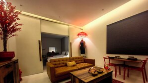 Uma sala de estar moderna com sofá, mesa de centro e área de jantar com cadeiras. Um espelho grande e uma luminária estão visíveis.