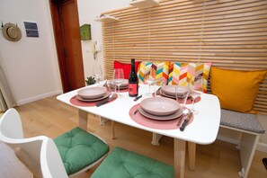 Comedor con una mesa blanca dispuesta para cuatro, incluyendo platos, cubiertos y copas de vino. Hay dos sillas con cojines verdes y un banco con un cojín amarillo. La habitación tiene persianas de madera y un cojín colorido en el banco.