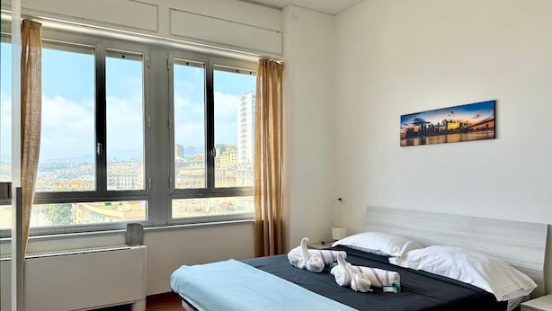 A hotel room with a bed, a nightstand, a wall-mounted painting, and a window with curtains. - TORRE CANTORE 69 (GENOVA OVEST/PORTO)   GENOVABB.IT (GENOVA)
