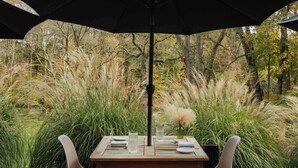 Un área de comedor al aire libre con una mesa para dos, rodeada de hierbas altas y árboles.