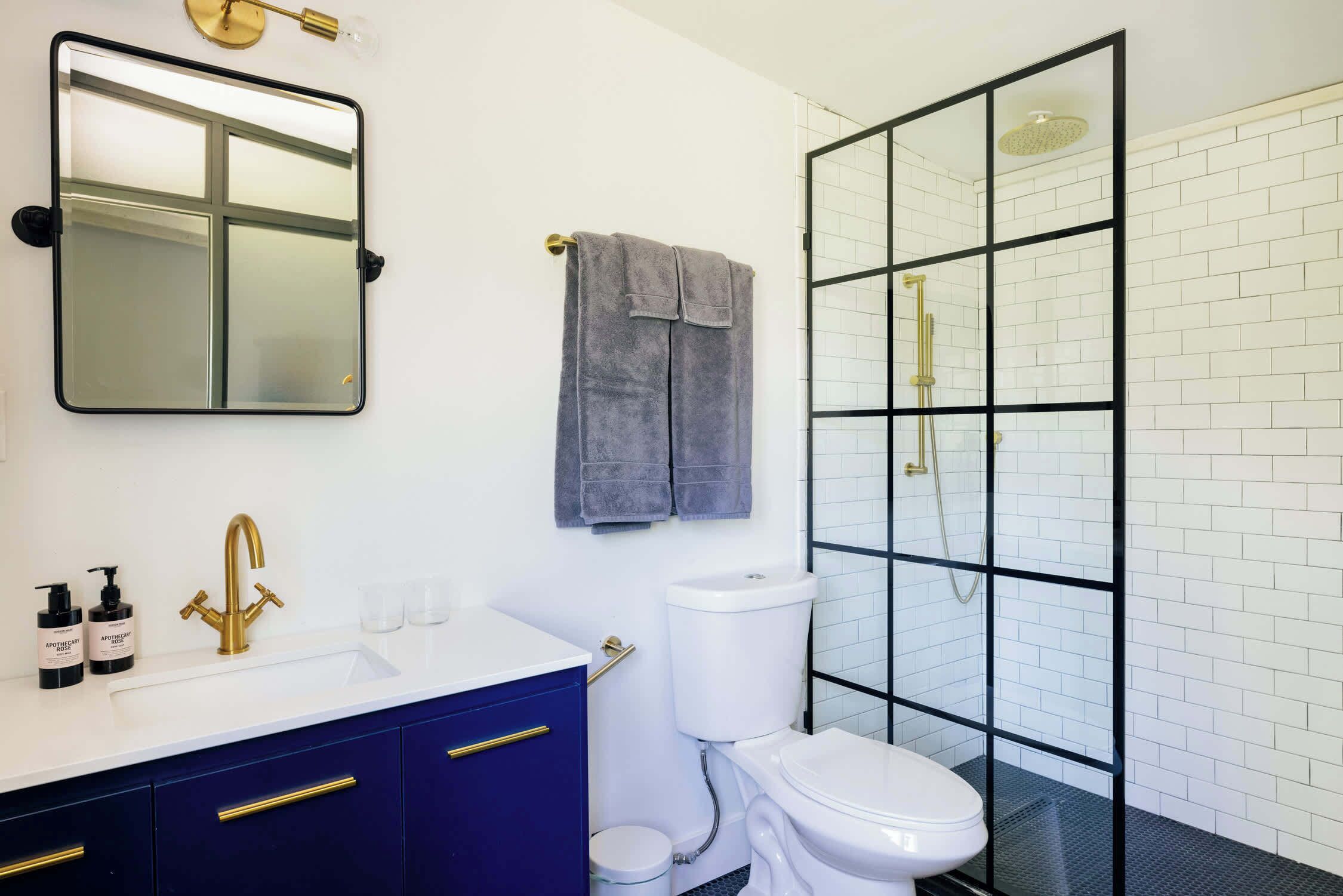 A modern bathroom with a white sink, blue cabinets, a gold faucet, a toilet, and a glass-enclosed shower.