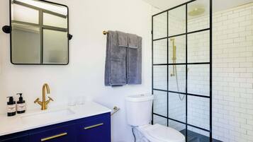A modern bathroom with a white sink, blue cabinets, a gold faucet, a toilet, and a glass-enclosed shower.