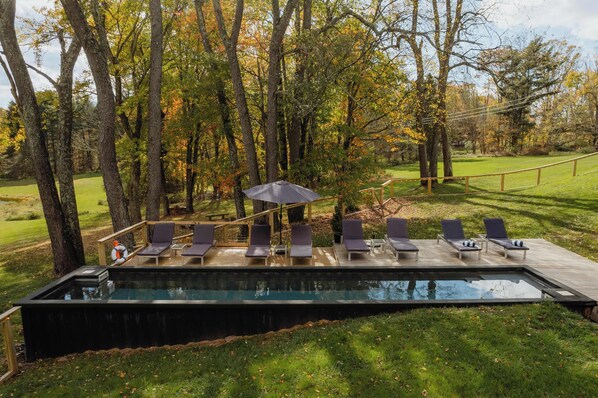 Piscina exterior moderna con sillones de descanso y una terraza de madera, rodeada de árboles y césped.