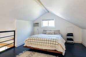 Un dormitorio con techo inclinado, una cama con colcha a cuadros, un ventanal, un equipo de aire acondicionado y un estante blanco pequeño.