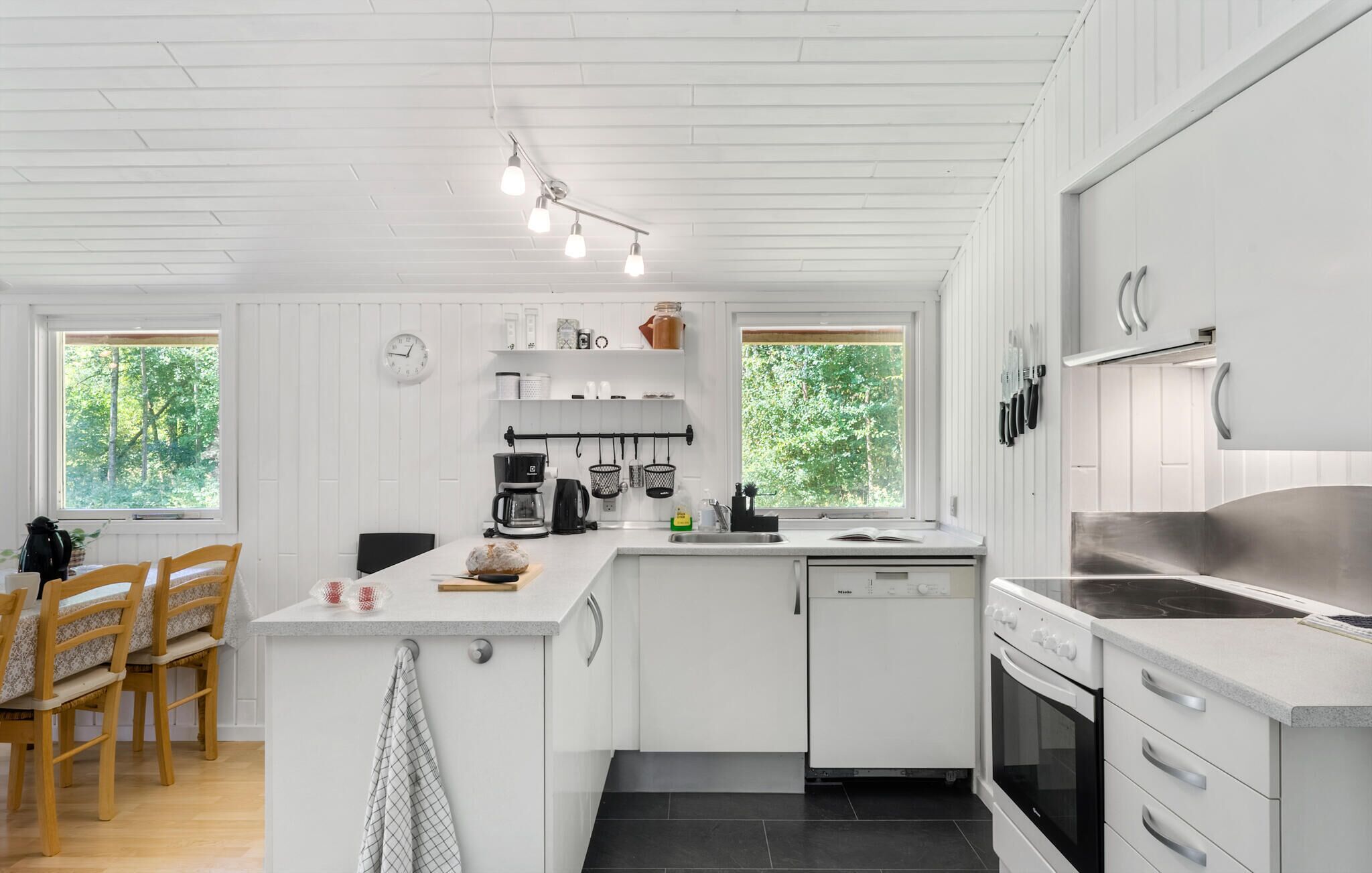 A modern kitchen with white cabinetry, a central island, a dishwasher, and a stove. There are two windows with a view of greenery outside.