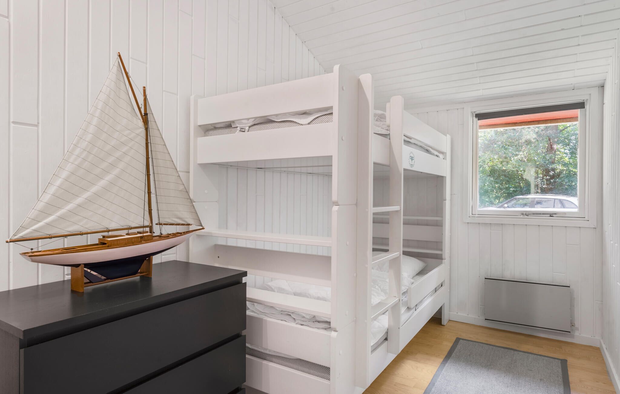 A room with a bunk bed, a small sailboat model, and a window with a view of trees and a car.