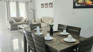 A dining area with a wooden table set for four, a ceiling fan, and a living area with a sofa and curtains. - Cozy 3-bedroom villa w/ AC, pool & unlimited beach access 10 mins from Ocho Rios (Saint Ann's Bay)