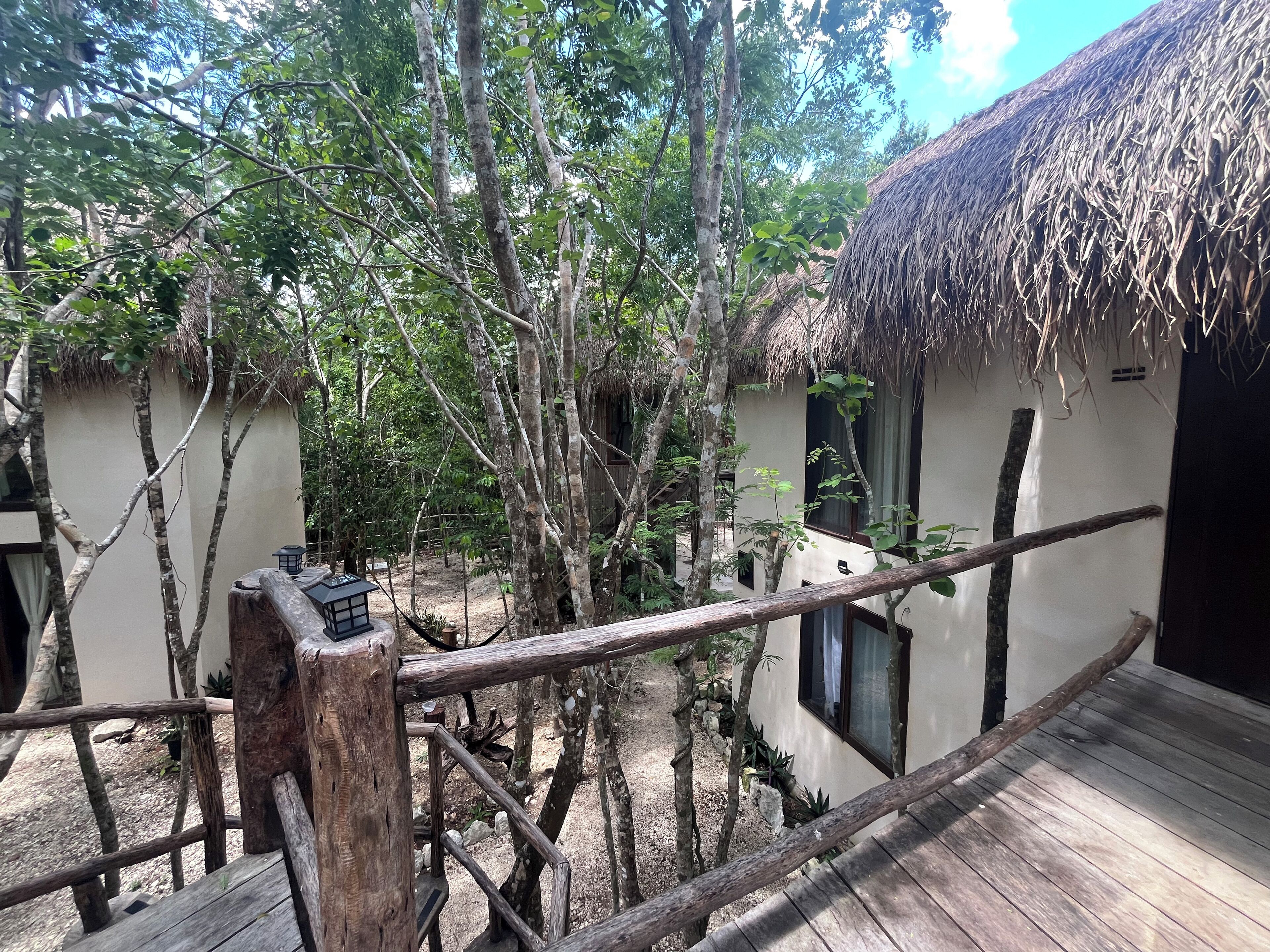 A wooden deck with a thatched roof structure, surrounded by trees and a white building with windows.