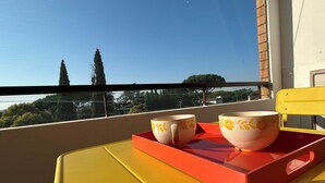 Un balcon avec deux tasses blanches ornées de motifs floraux jaunes, posées sur un plateau rouge, d’où l’on peut admirer un paysage urbain avec deux grands immeubles et des arbres.