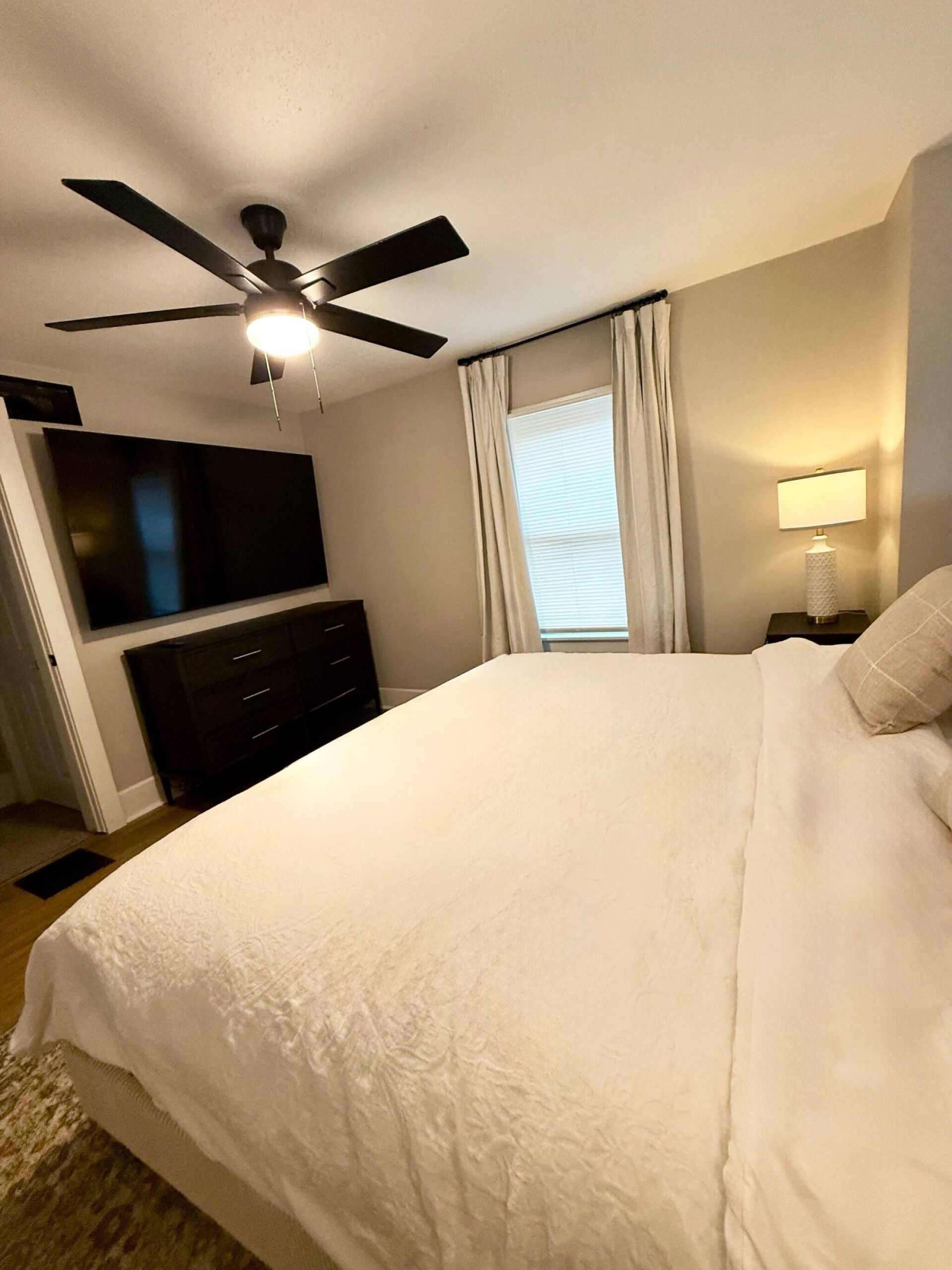 Une chambre à coucher comprenant un lit, un ventilateur de plafond, une télévision, une commode, une fenêtre avec des rideaux et une lampe de chevet.