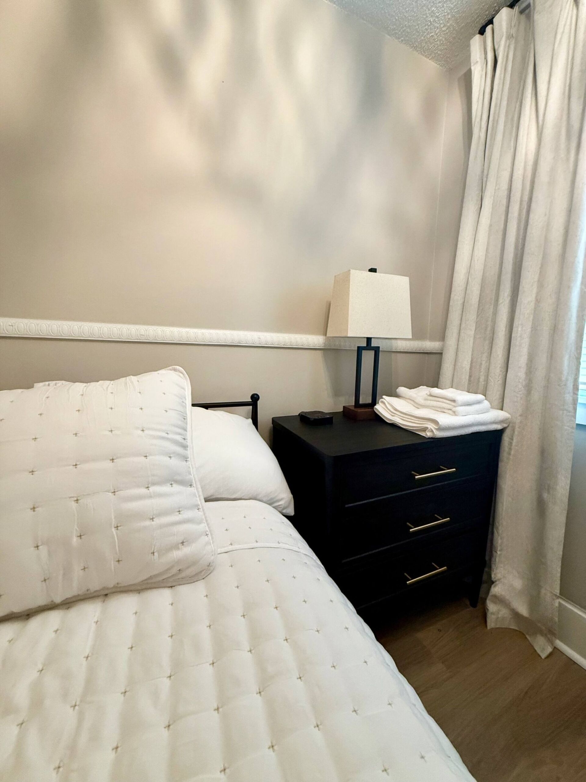 A neatly made bed with white bedding, a bedside table with a lamp, and a nightstand with folded towels.