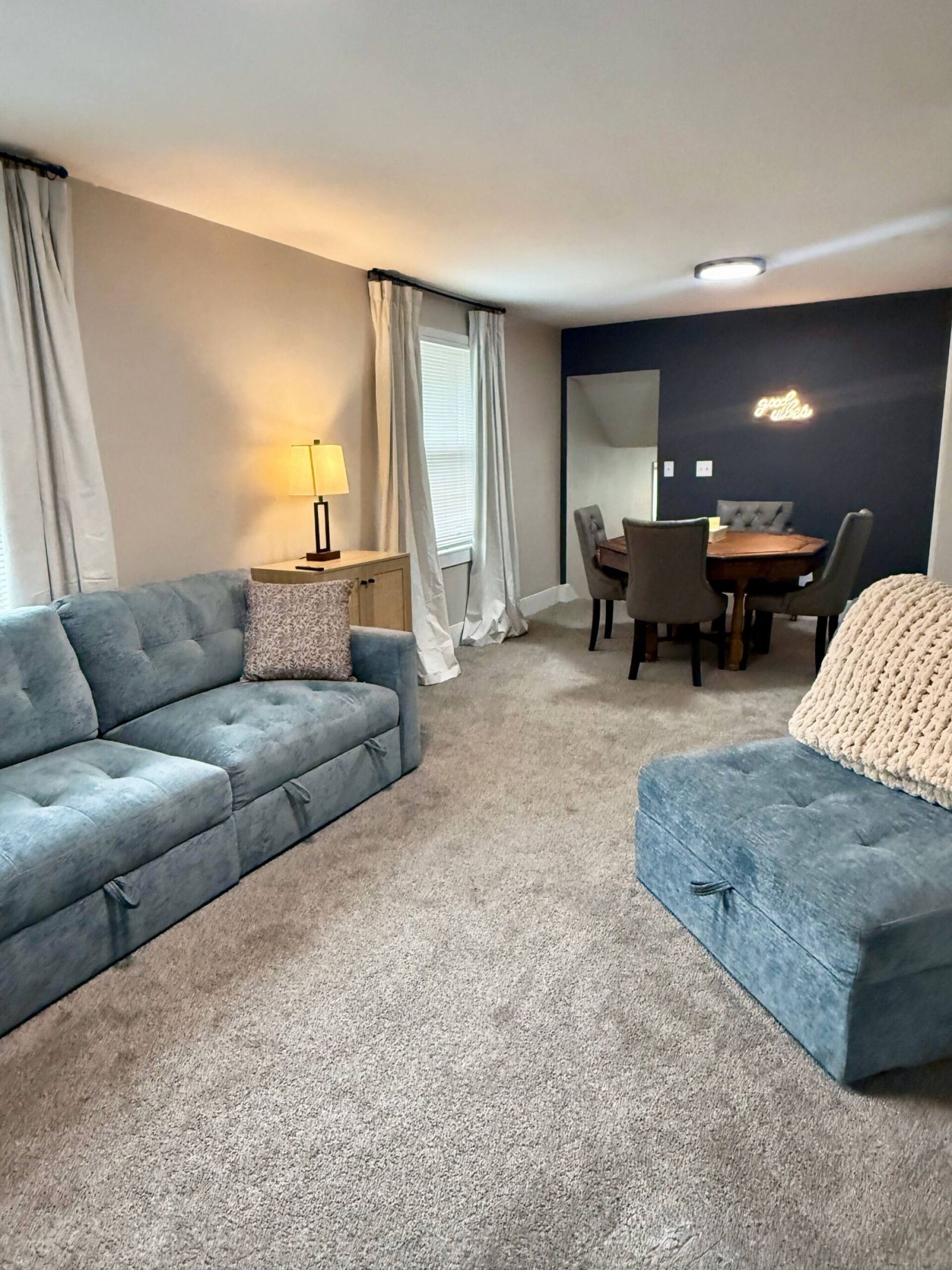 A living room with a sofa, ottoman, dining table, chairs, lamp, and curtains.