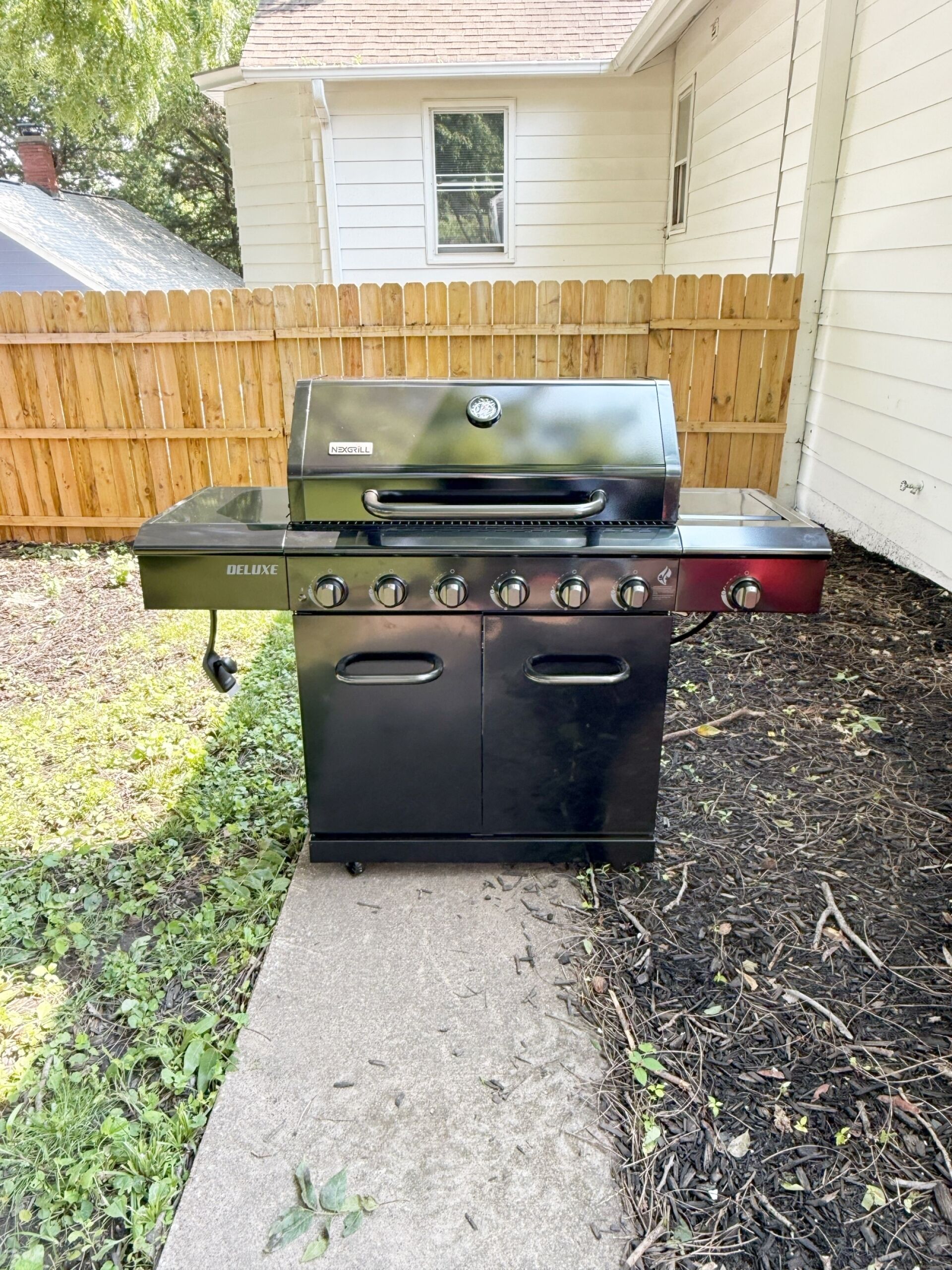 En sort udendørs grill med flere brændere og en sidebrænder, placeret på en betonplade i en baghave.