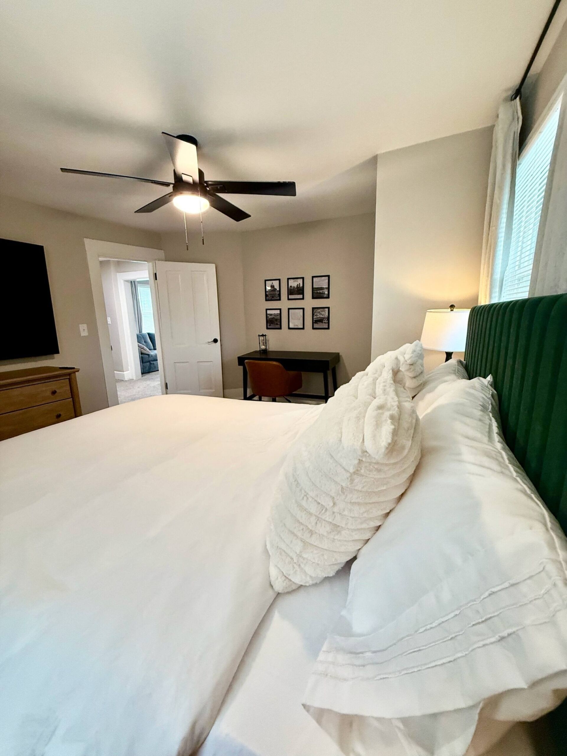 A hotel room with a bed, a desk, a chair, a ceiling fan, and a door leading to another room.