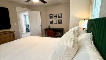 A hotel room with a bed, a desk, a chair, a ceiling fan, and a door leading to another room.