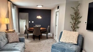 A modern living room with a grey sofa, a blue ottoman, a dining table with chairs, a wall-mounted TV, and a ceiling light.