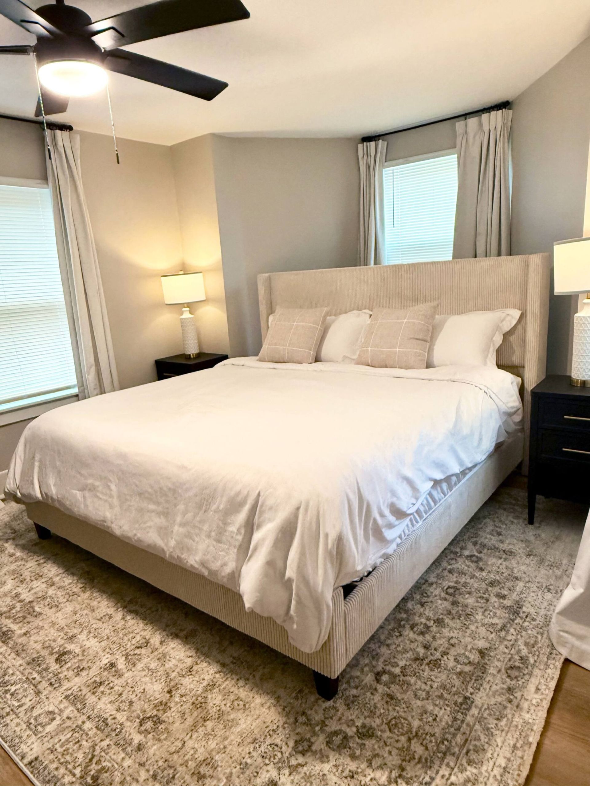 Une chambre à coucher avec un lit, des tables de chevet, un ventilateur de plafond et une fenêtre avec des rideaux.