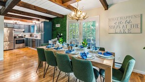 A dining area with a wooden table set for six, surrounded by green upholstered chairs. A chandelier hangs from the ceiling, and a sign on the wall reads "What happens at the lake stays at the lake." - Night Golf, Fire Pit, Game Room, Hot Tub, Theater room, Lake Access Fun house! (East Stroudsburg)