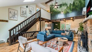 A spacious living room with a blue sectional sofa, a coffee table, and a fireplace. There is a staircase with wooden railings leading to an upper level. - Night Golf, Fire Pit, Game Room, Hot Tub, Theater room, Lake Access Fun house! (East Stroudsburg)