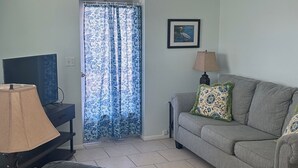 A living room with a grey sofa, a black armchair, a TV on a stand, a lamp, and a door with blue floral curtains. - Emerald Palms Unit 2 (Englewood)