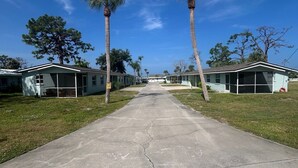 A row of single-story mobile homes with a concrete driveway, surrounded by grass and palm trees, under a clear blue sky. - Emerald Palms Unit 2 (Englewood)