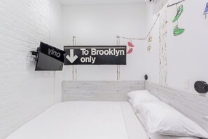 Una habitación con una cama, una pared de ladrillo blanco, un letrero que indica Brooklyn y un televisor instalado en la pared.