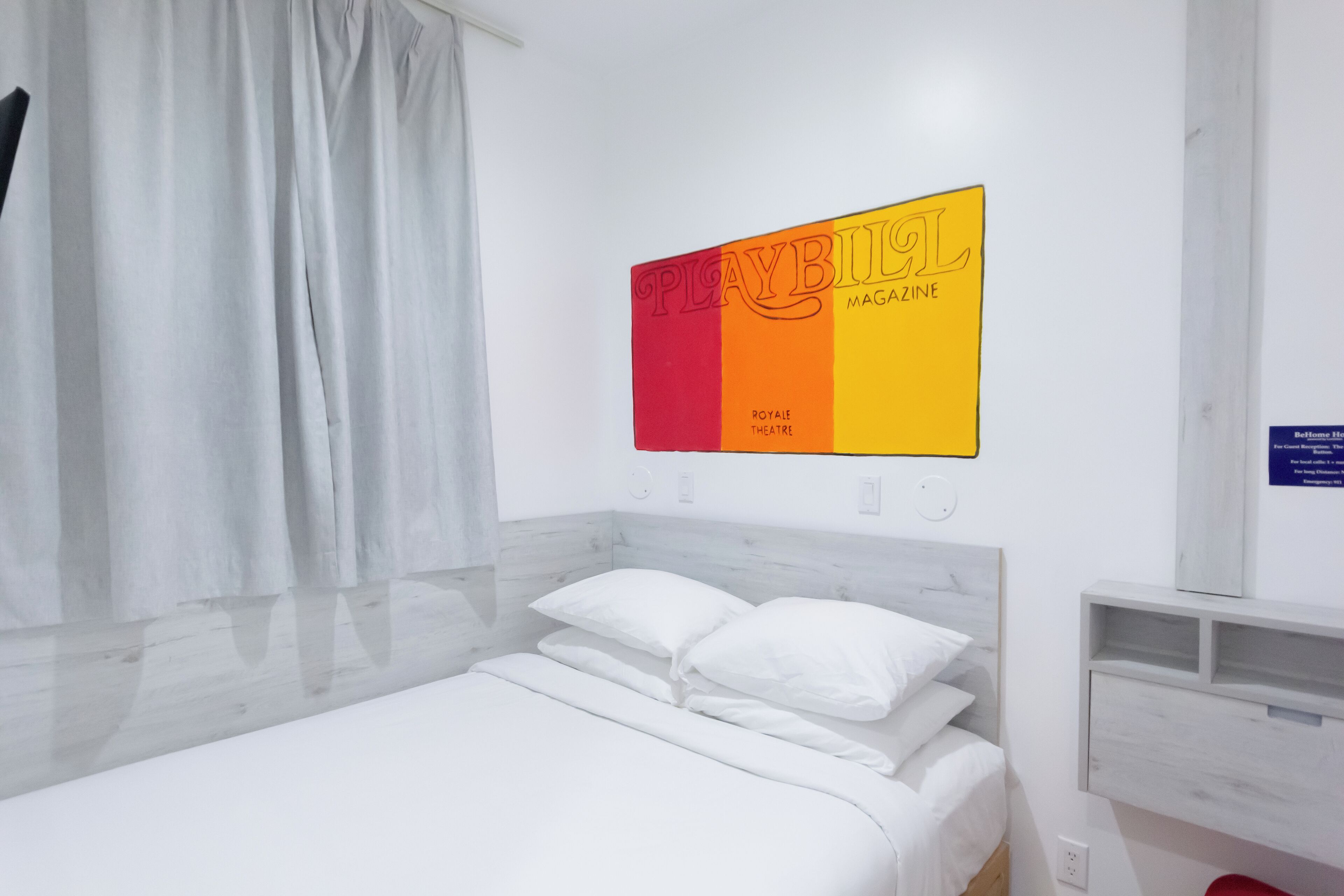 A modern hotel room with a bed, white curtains, a wall-mounted artwork, and a small shelf.