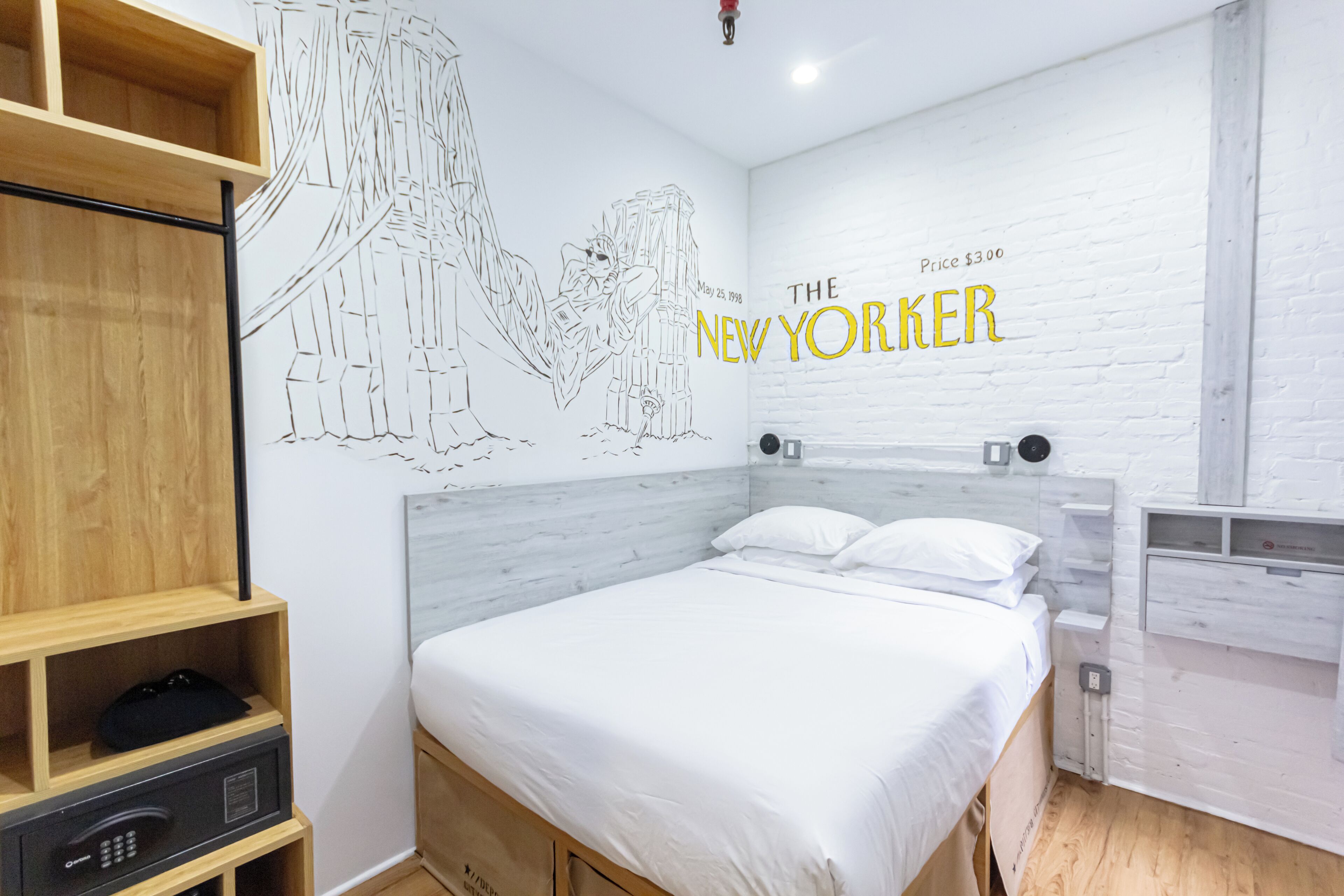 Une chambre d’hôtel avec un lit, une étagère en bois, une fresque murale et un mur en briques blanches sur lequel est inscrit le texte « THE NEW YORKER ».