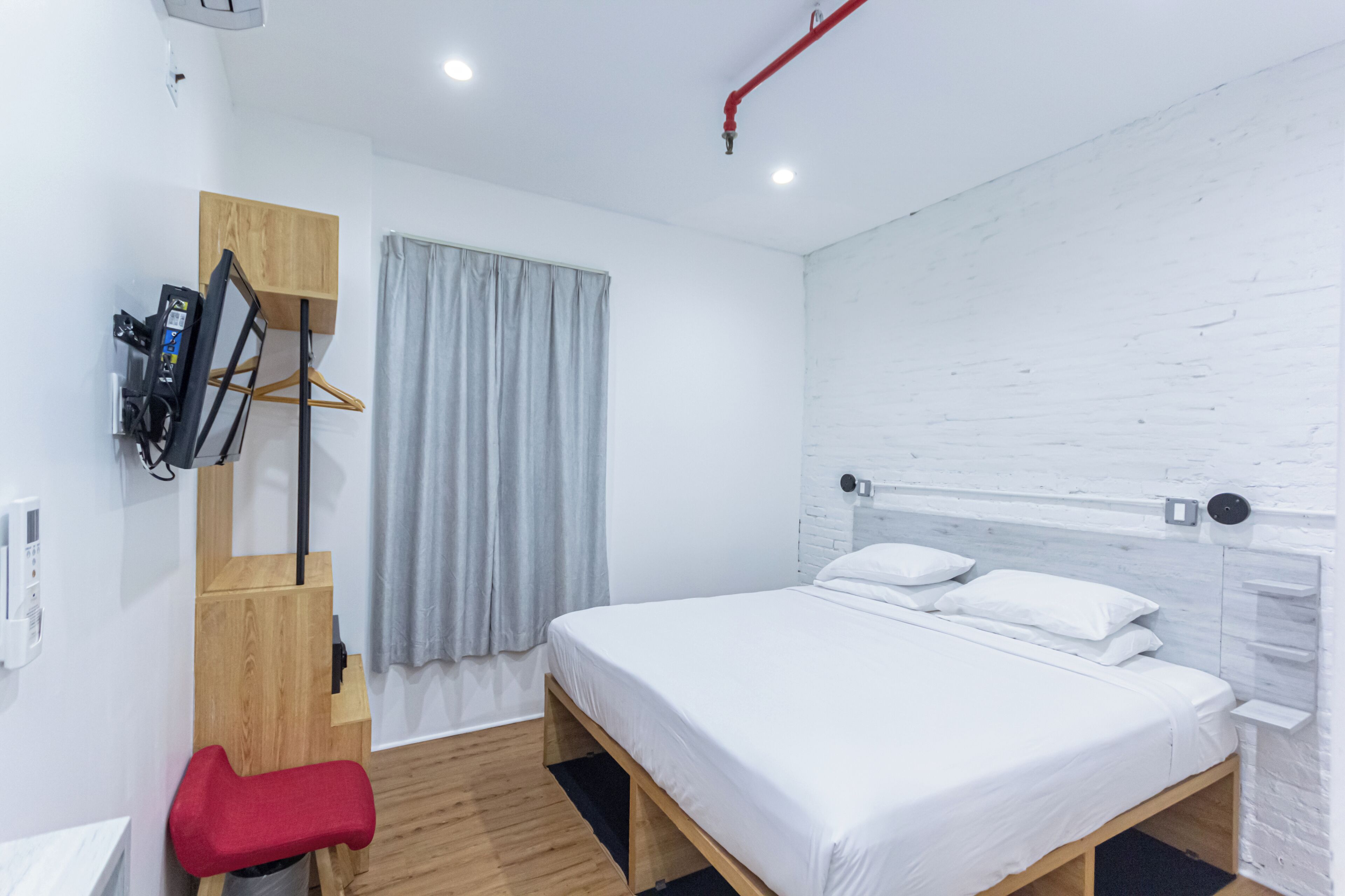 Una habitación de hotel moderna con un cabecero de madera, una silla roja, un televisor y una cortina gris.