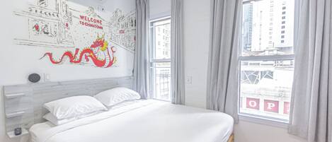 Une chambre d’hôtel moderne avec un grand lit, une vue sur la ville par la fenêtre et une fresque murale représentant un dragon ainsi que le texte « BIENVENUE À CHINATOWN ».