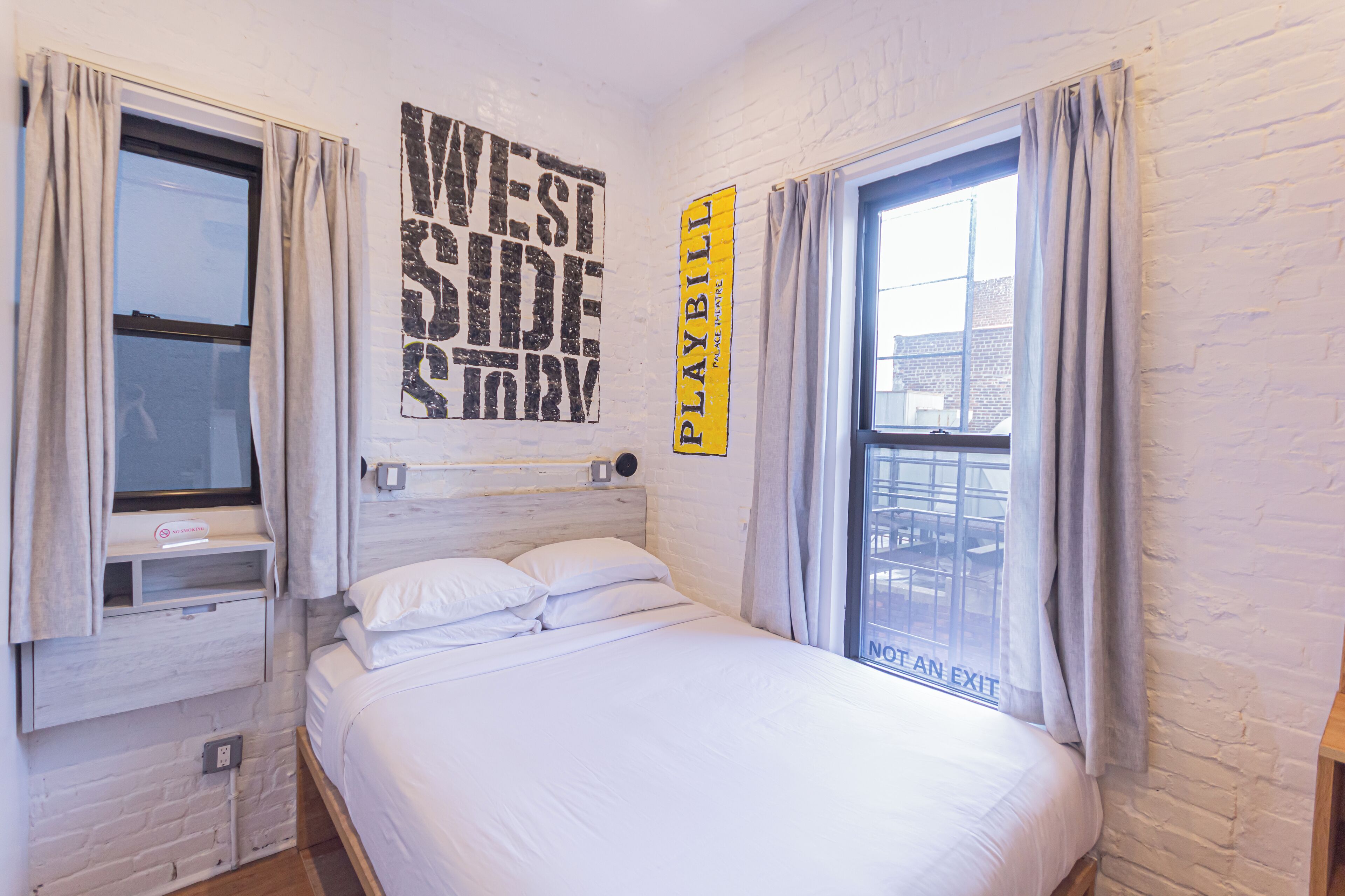 Ein Zimmer mit einem Einzelbett, einer weißen Backsteinwand, einem Fenster mit Vorhängen und einem 'West Side Story'-Poster.