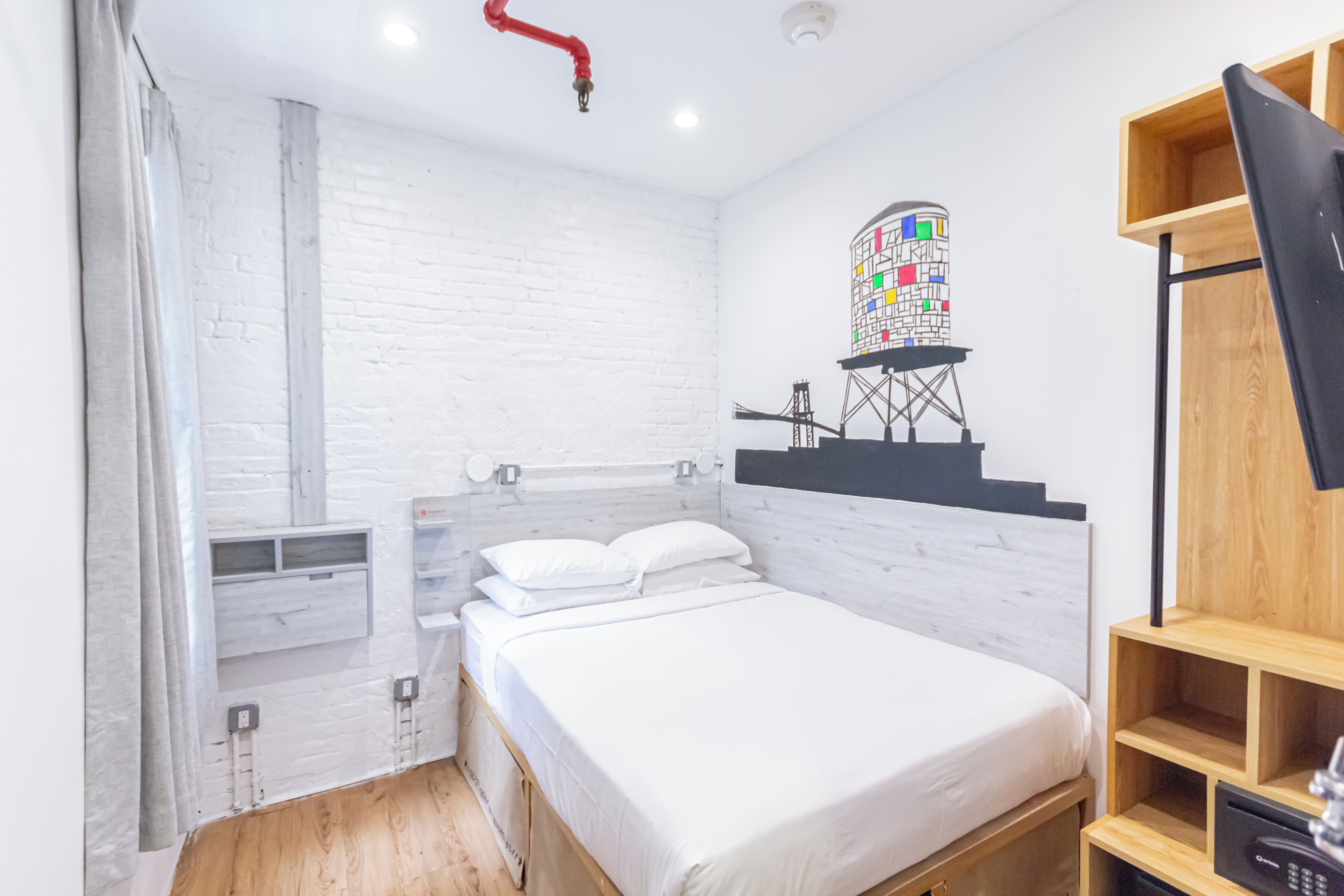 A modern bedroom with a white brick wall, a bed with white bedding, a wooden floor, a small window with a colorful stained glass design, and a wooden shelf unit.