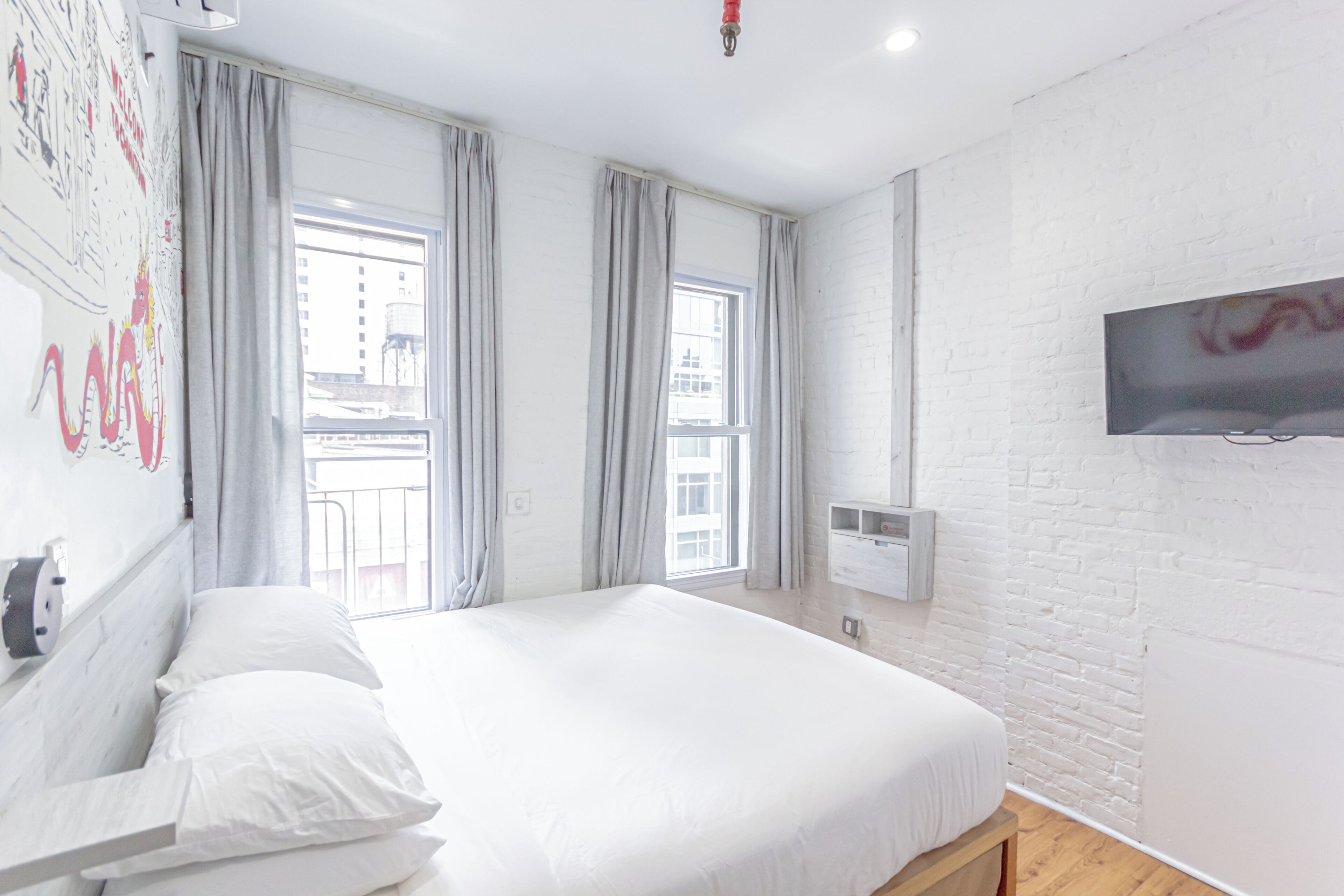 A modern hotel room with a large bed, two windows with curtains, a mounted TV, and a white brick wall.