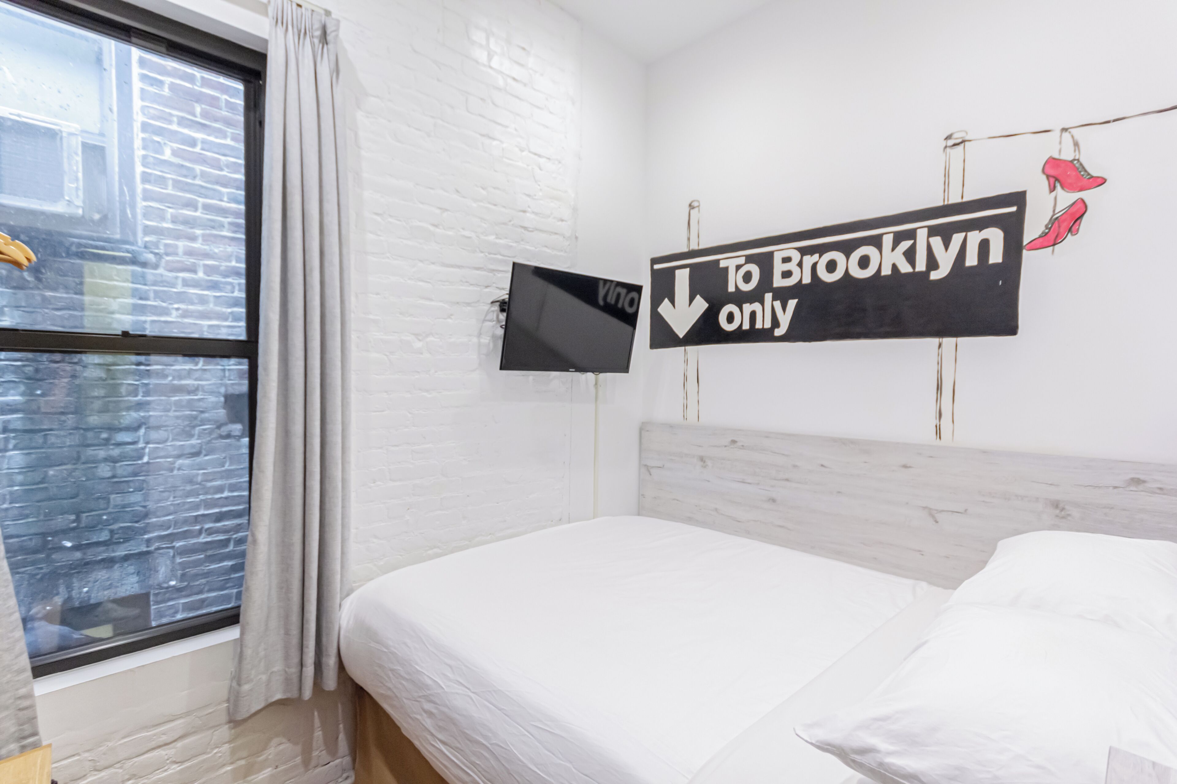 A hotel room with a bed, a window with curtains, a TV mounted on the wall, and a sign indicating the room is for 'Brooklyn only'.