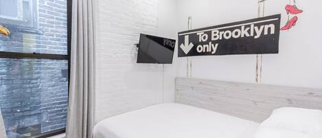 A hotel room with a bed, a window with curtains, a TV mounted on the wall, and a sign indicating the room is for 'Brooklyn only'.