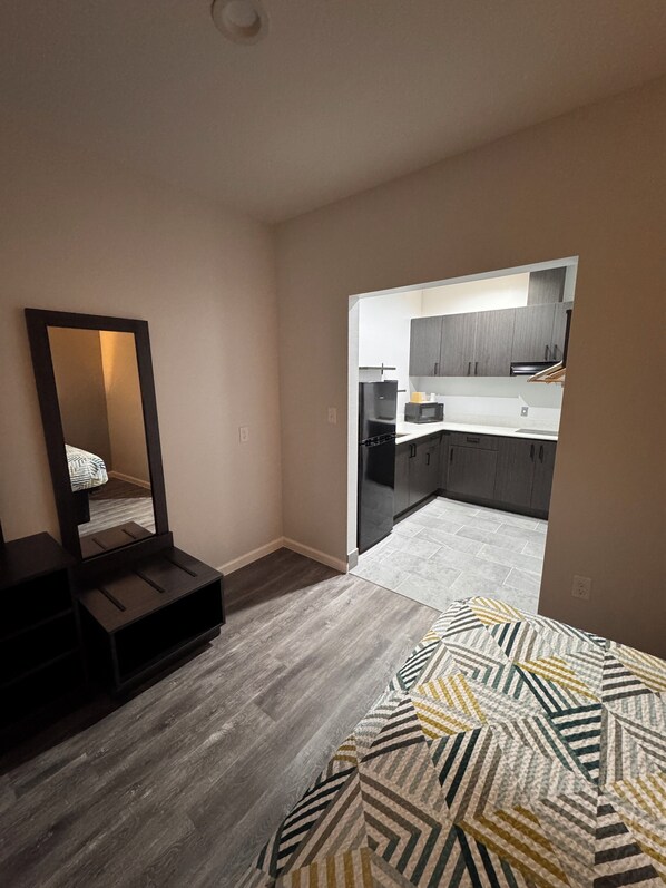 Un dormitorio con cama, cómoda y espejo. Se ve una cocina a través de una puerta abierta.