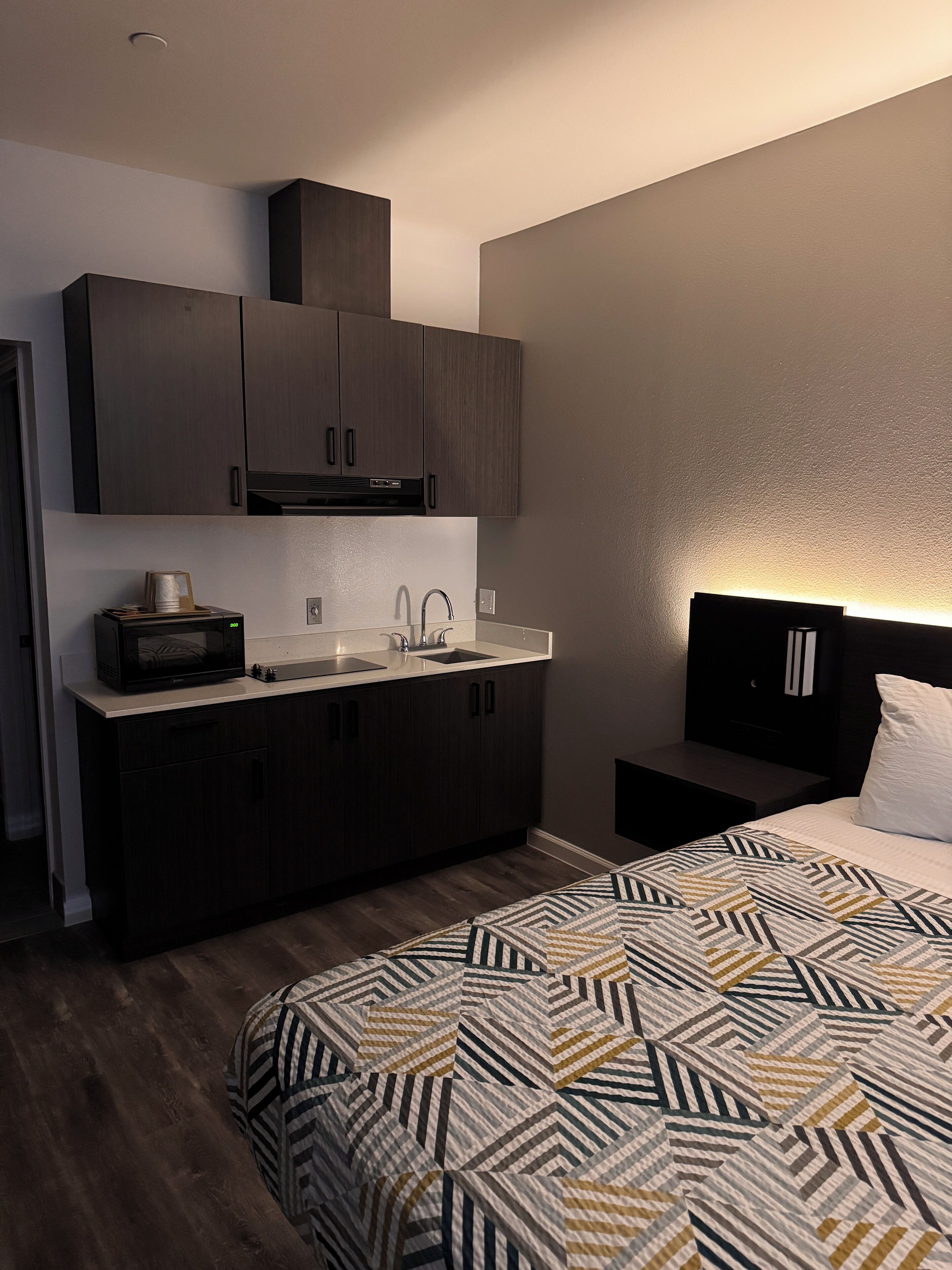 A compact hotel room with a kitchenette, a bed with a patterned bedspread, and a nightstand with a lamp.